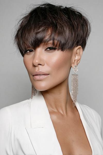 A woman with a dark brown undercut pixie hairstyle featuring textured, voluminous layers on top and short sides, styled with a full, wispy fringe covering the forehead.