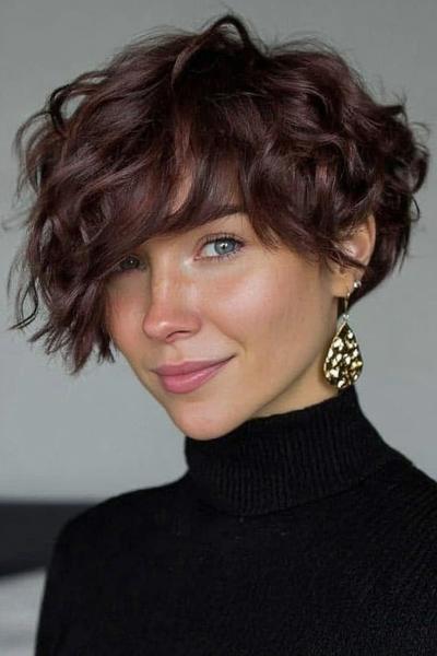 A woman with a dark, curly undercut pixie haircut featuring longer, tousled layers on top and shorter sides, styled with a side fringe.