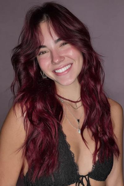 A woman with long, wavy deep red hair featuring soft curtain bangs, styled in loose layers.