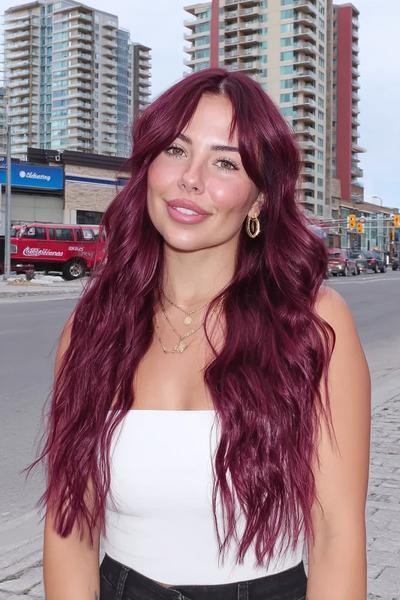 A woman with long, wavy deep red hair styled in loose layers and curtain fringe, standing outdoors in an urban setting.