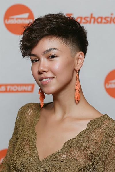A young woman with thick, wavy hair wears an undercut pixie hairstyle, featuring closely shaved sides and back with longer, textured layers on top.