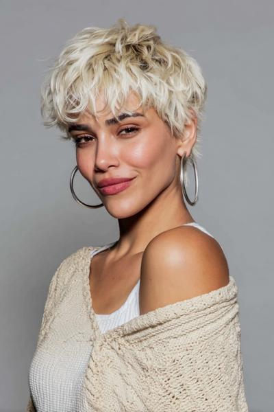 A woman with platinum blonde, textured undercut pixie hair styled in soft waves, wearing large hoop earrings and a cream knit top.