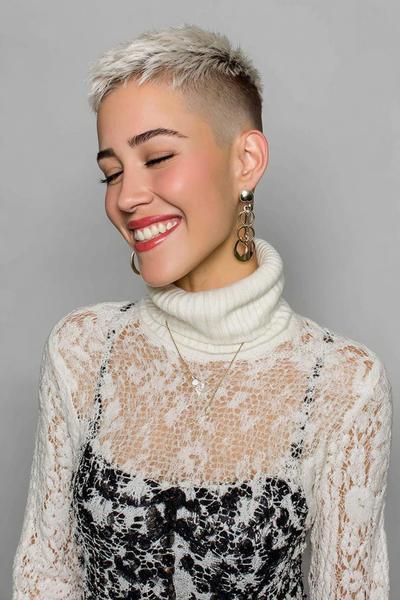 A woman with a platinum blonde, closely cropped undercut pixie hairstyle, featuring longer textured hair on top and shaved sides, is smiling with her eyes closed.