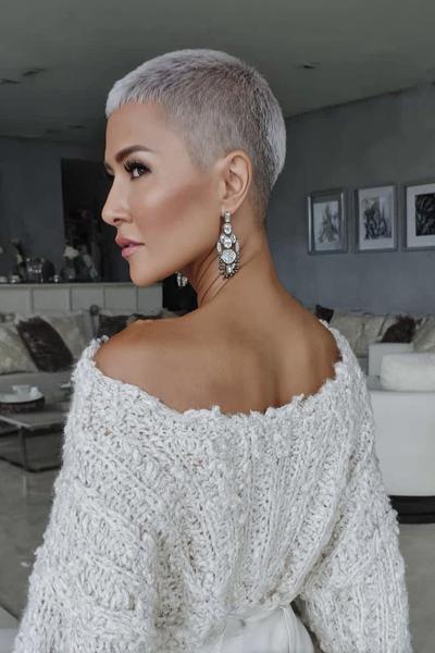 A woman with a platinum blonde undercut pixie hairstyle, featuring very short sides and back with slightly longer hair on top, is pictured indoors wearing a chunky knit off-the-shoulder jumper and statement earrings.