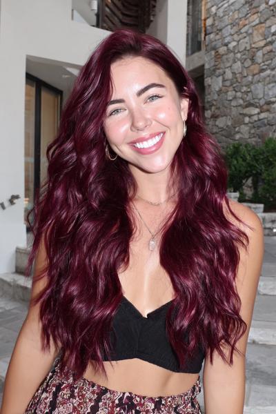 A smiling woman with long, wavy burgundy red hair is wearing a black crop top and patterned trousers, standing outdoors in front of a stone building.