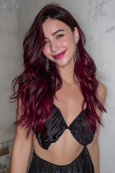 A woman with long, wavy, rich burgundy red hair is styled in loose waves, wearing a black top and drop earrings.