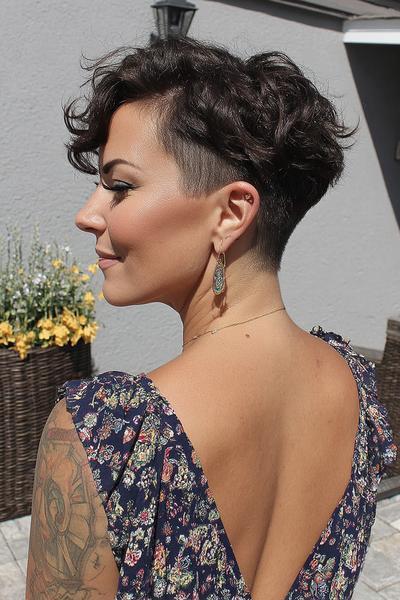 A woman with dark, curly hair is sporting a short pixie cut with a dramatic undercut on the sides and back, while the top remains voluminous and textured.