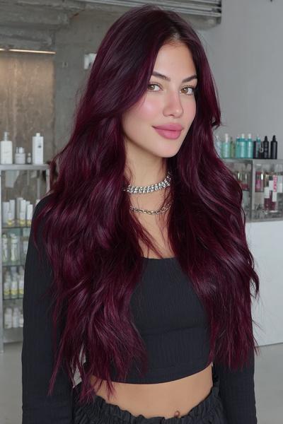 A woman with long, wavy, deep burgundy red hair is wearing a black crop top and layered necklaces in a modern salon setting.